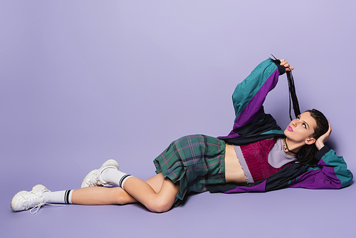 pensive woman in vintage clothes and white sneakers holding braided dreadlocks while lying on purple background
