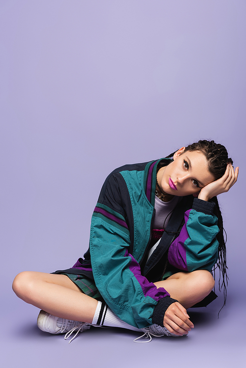 thoughtful woman in nineties style jacket sitting with crossed legs and looking at camera on purple background