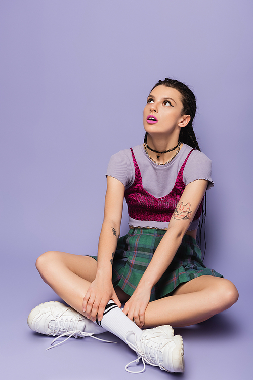 amazed woman in plaid skirt and white sneakers sitting with crossed legs and looking away on purple background