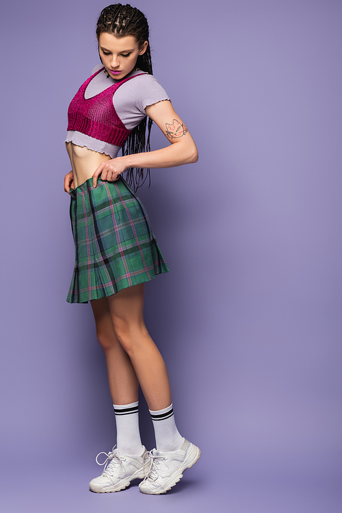 full length of woman in crop top and white sneakers adjusting checkered skirt on purple background