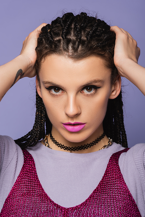 portrait of pretty woman in makeup touching braided hair while looking at camera isolated on purple