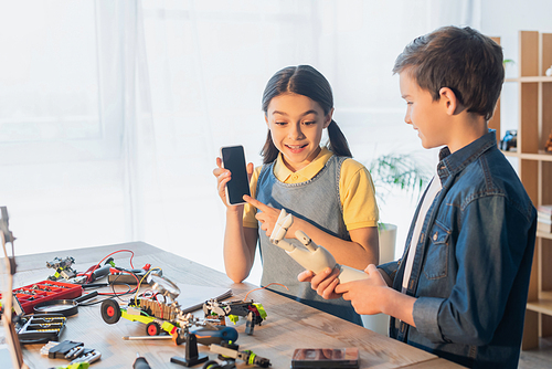 amazed girl pointing at smartphone with blank screen near boy holding robotics hand at home