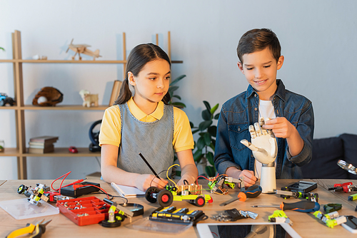 girl writing in notebook while boy holding robotics hand near mechanical parts and blurred digital tablet