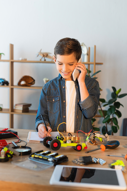 smiling boy talking on smartphone and writing in notebook near mechanical parts and blurred digital tablet