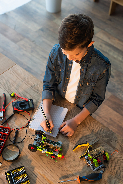 top view of boy writing in notebook near tools and details of robotics model on table