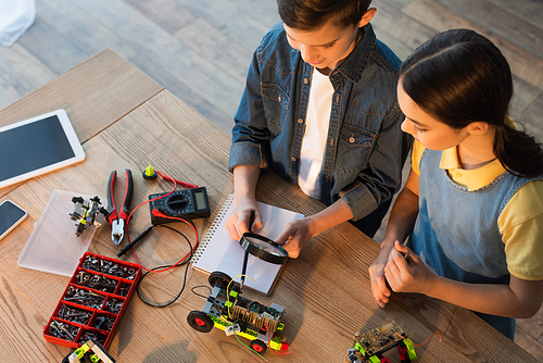 high angle view of boy holding magnifier near notebook and pointing with pencil at robotics model