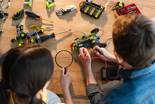 top view of boy with multimeter and girl with magnifying glass assembling robotics model at home