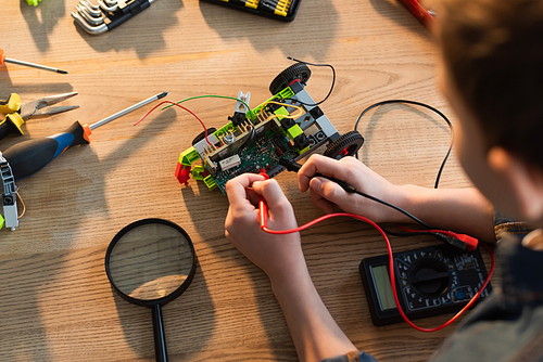 top view of cropped boy with multimeter measuring voltage of robotics model near magnifier