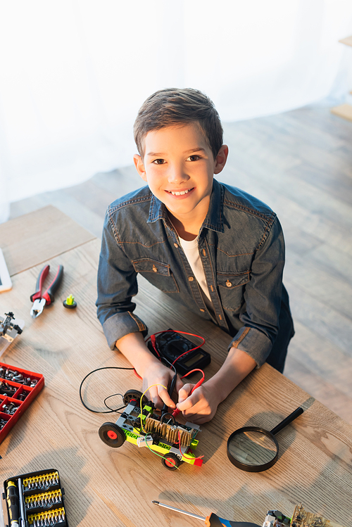 high angle view of smiling boy with multimeter measuring voltage of robotics model near magnifier