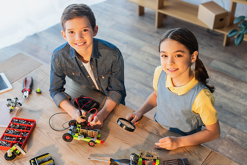 high angle view of kids with multimeter and magnifier assembling robotics model and smiling at camera