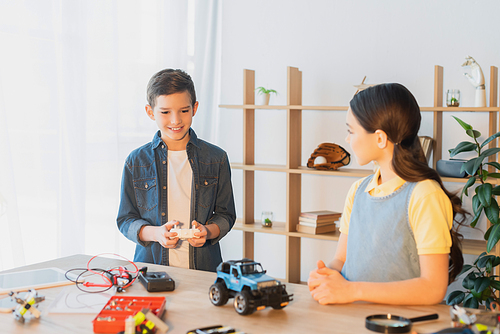 happy boy holding remote controller near handmade car model and girl at home