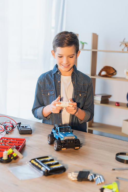 happy boy holding remote controller near handmade car model on table at home