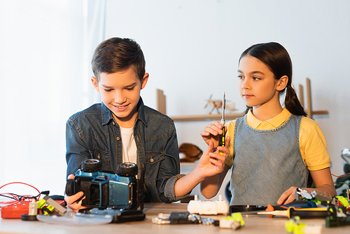 preteen girl holding screwdriver near friend looking at handmade car model at home