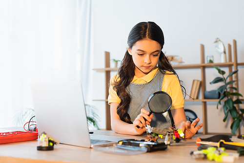 brunette girl with magnifying glass near laptop and blurred parts of robotics model