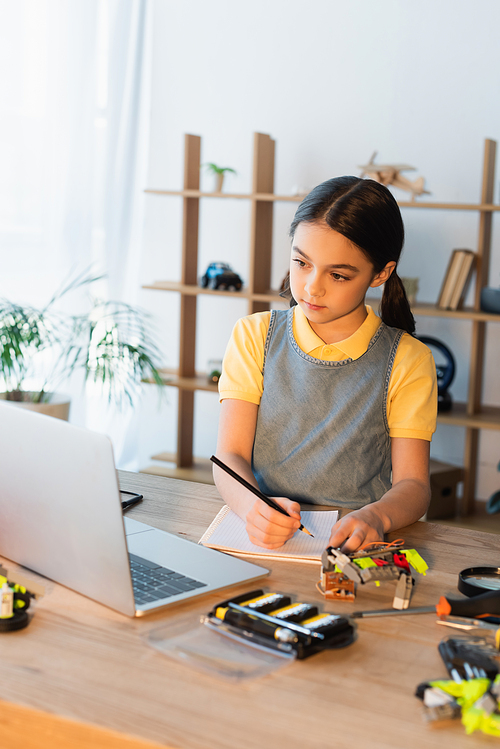 preteen girl looking at laptop and writing in notebook near details of robotics model