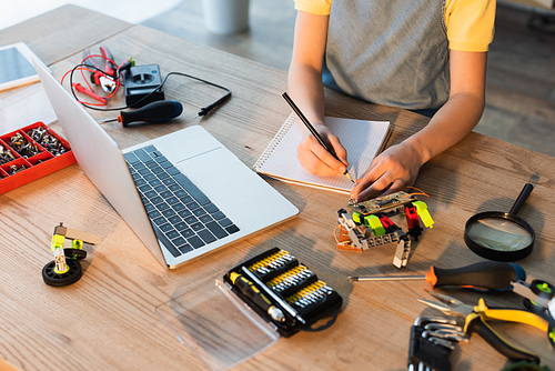 high angle view of cropped girl writing in notebook near laptop and mechanical parts of robotics model