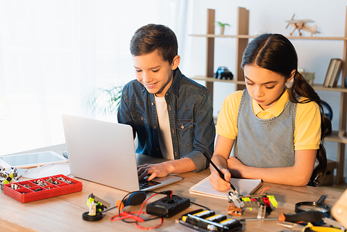 happy boy using laptop while girl writing in notebook near details of robotics model