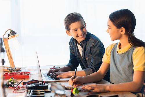 smiling boy using laptop while looking at girl writing near blurred parts of robotics model