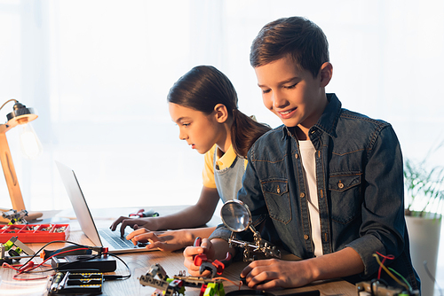 smiling boy with magnifier assembling robotics model near girl using laptop