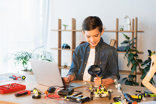 smiling boy holding magnifying glass near parts of robotics model and laptop