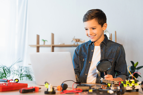 smiling boy with magnifier looking at laptop near blurred mechanical parts on table at home