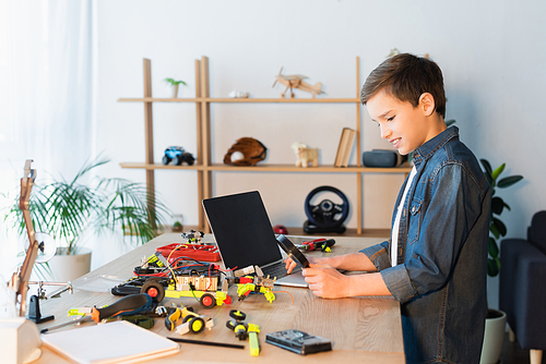 side view of kid with magnifying glass near details of robotics model and laptop with blank screen