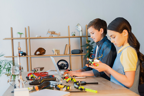 preteen boy using laptop near girl holding mechanical part of robotics model