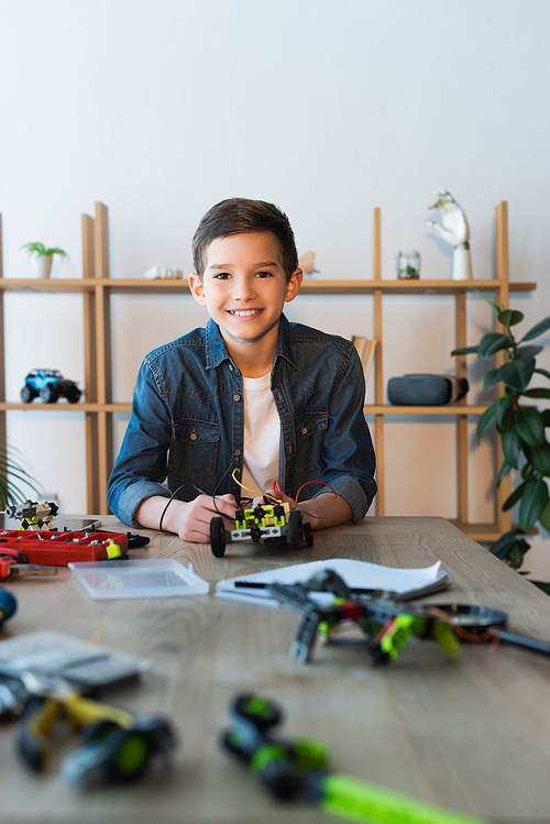 cheerful boy looking at camera near mechanical details of robotics model on table at home