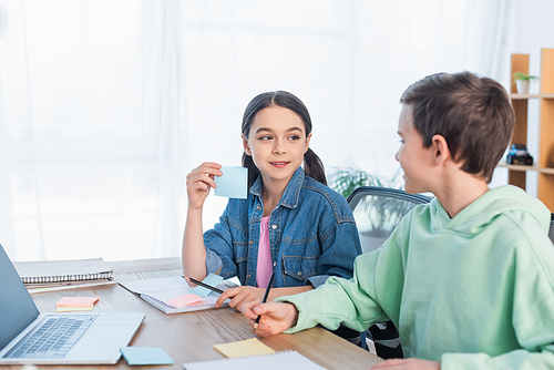 smiling girl holding empty sticky note near blurred boy and laptop on desk