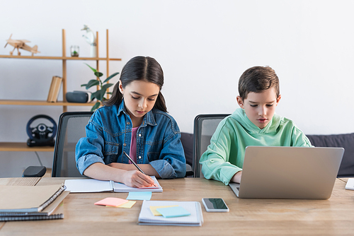 boy using laptop and girl writing in notebook near smartphone with blank screen on desk at home