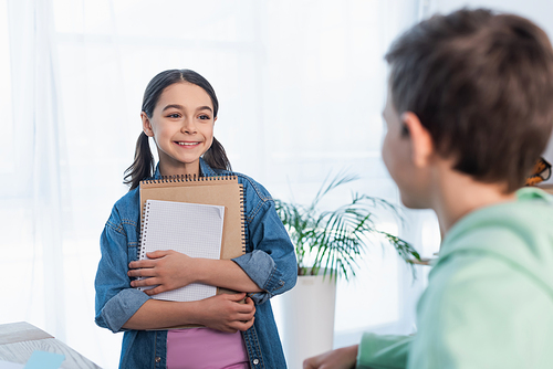 cheerful girl holding notebooks and looking at blurred boy at home