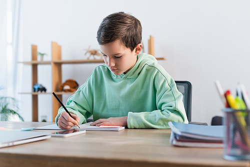 focused boy doing homework and writing in copybook near blurred mobile phone