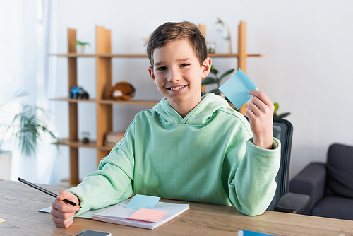 happy boy holding empty sticky note and pencil near copybook on desk at home