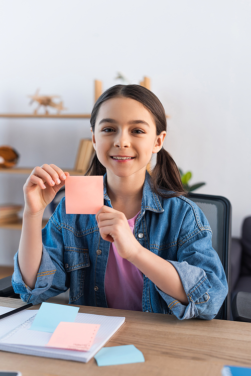 cheerful girl showing empty sticky note and looking at camera at home