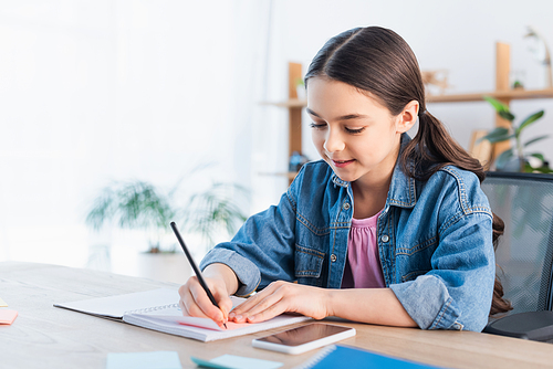 smiling brunette girl writing in notebook while doing homework near cellphone with blank screen