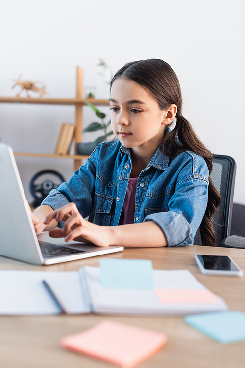 focused schoolgirl typing on laptop while learning at home near smartphone with blank screen