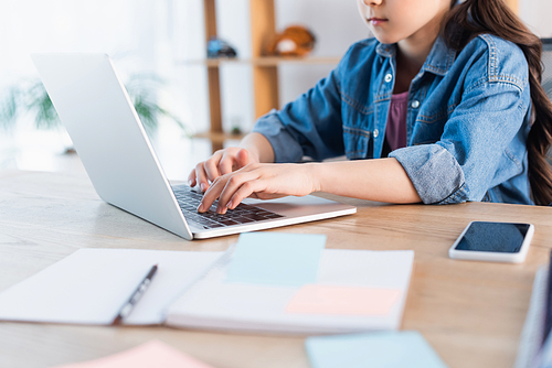 cropped view of girl typing on laptop near blurred copybook while studying at home