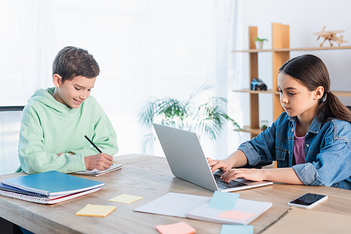 positive boy writing in notebook and girl typing on laptop while studying at home