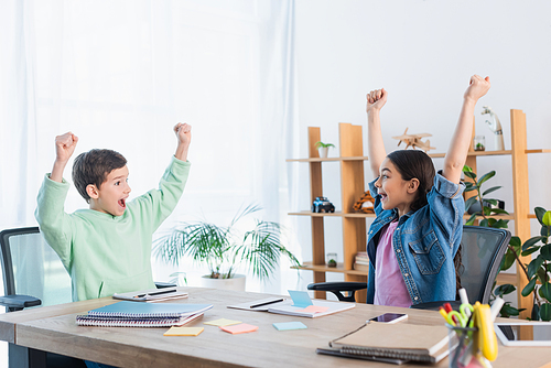 excited kids showing win gesture near notebooks and paper notes on table at home