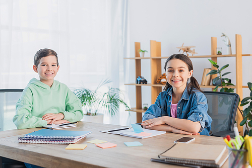 cheerful kids sitting at desk near notebooks and sticky notes while smiling at camera