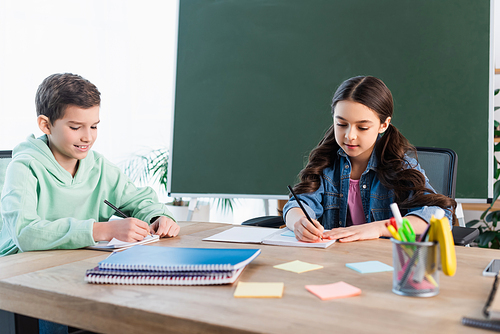 smiling boy writing in notebook near girl and chalkboard in school