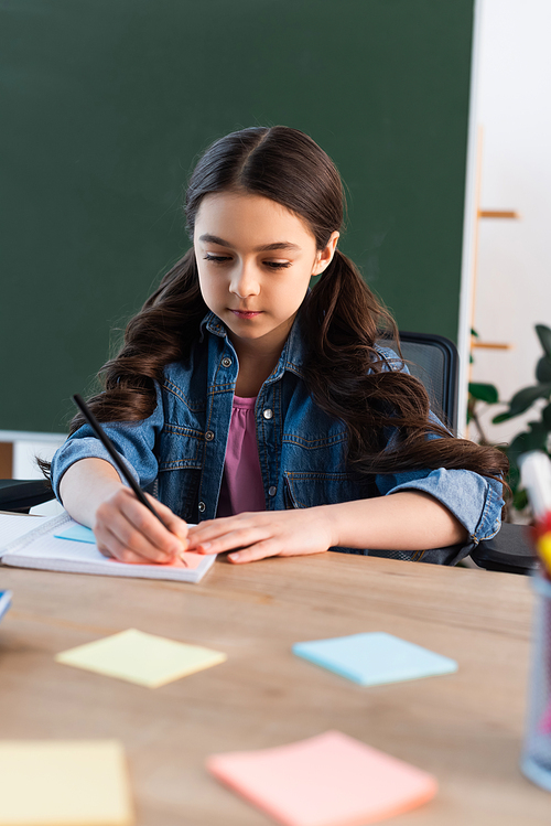 brunette girl writing in notebook near blurred sticky notes and chalkboard in school