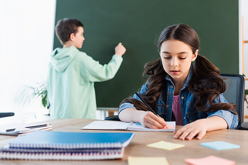 brunette schoolgirl writing in notebook near boy at chalkboard on background