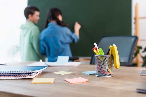 selective focus of school desk with notebooks and sticky notes near blurred classmates writing on chalkboard