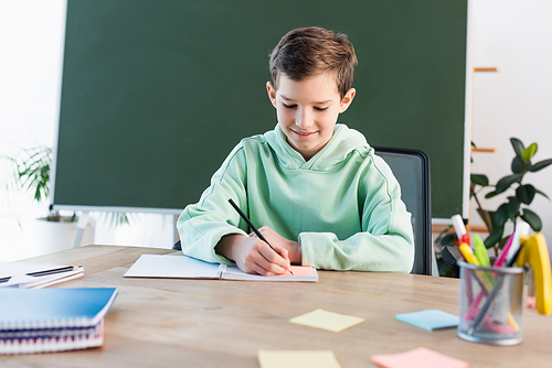 smiling boy writing in notebook near chalkboard and blurred sticky notes on school desk