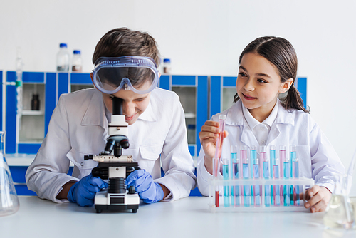smiling girl holding test tube near friend looking into microscope during chemical experiment