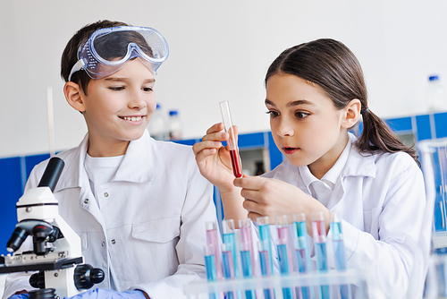 amazed girl holding test tube with red liquid near smiling boy and microscope in lab