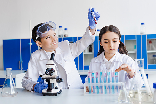 cheerful boy holding test tube with red liquid near microscope and serious girl in lab