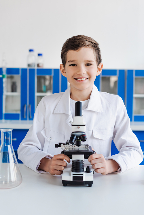 happy preteen kid in white coat looking at camera near microscope in laboratory