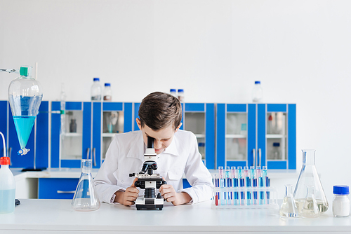 boy in white coat looking into microscope near test tubes and flasks in lab